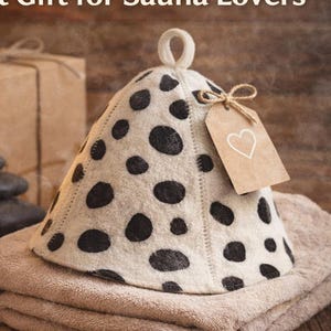 Peut inclure: Un chapeau de sauna crème à pois noirs est posé sur une serviette beige pliée. Une petite étiquette avec un cœur est attachée. Le texte "Perfect Gift for Sauna Lovers" est en haut. Le chapeau est en laine respirante.