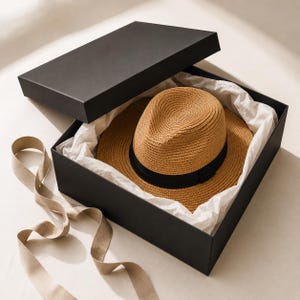 May include: A tan straw fedora hat with a black ribbon band, nestled inside a black rectangular gift box lined with white tissue paper. The box lid is partially open, and a long, light beige ribbon extends from the base of the box.