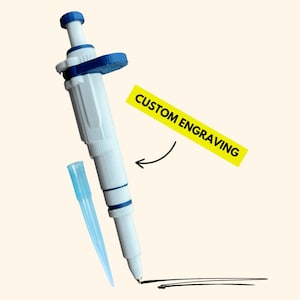 May include: A white and blue laboratory micropipette with a plunger mechanism and a detachable light blue tip. A yellow rectangular label with the black text CUSTOM ENGRAVING points to the body of the pipette, which is drawing a black line.