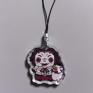 May include: A clear acrylic charm featuring a cartoon character with a chainsaw and a snowman. The character has dark curly hair, a white shirt, and a red skirt. The charm is attached to a black cord.