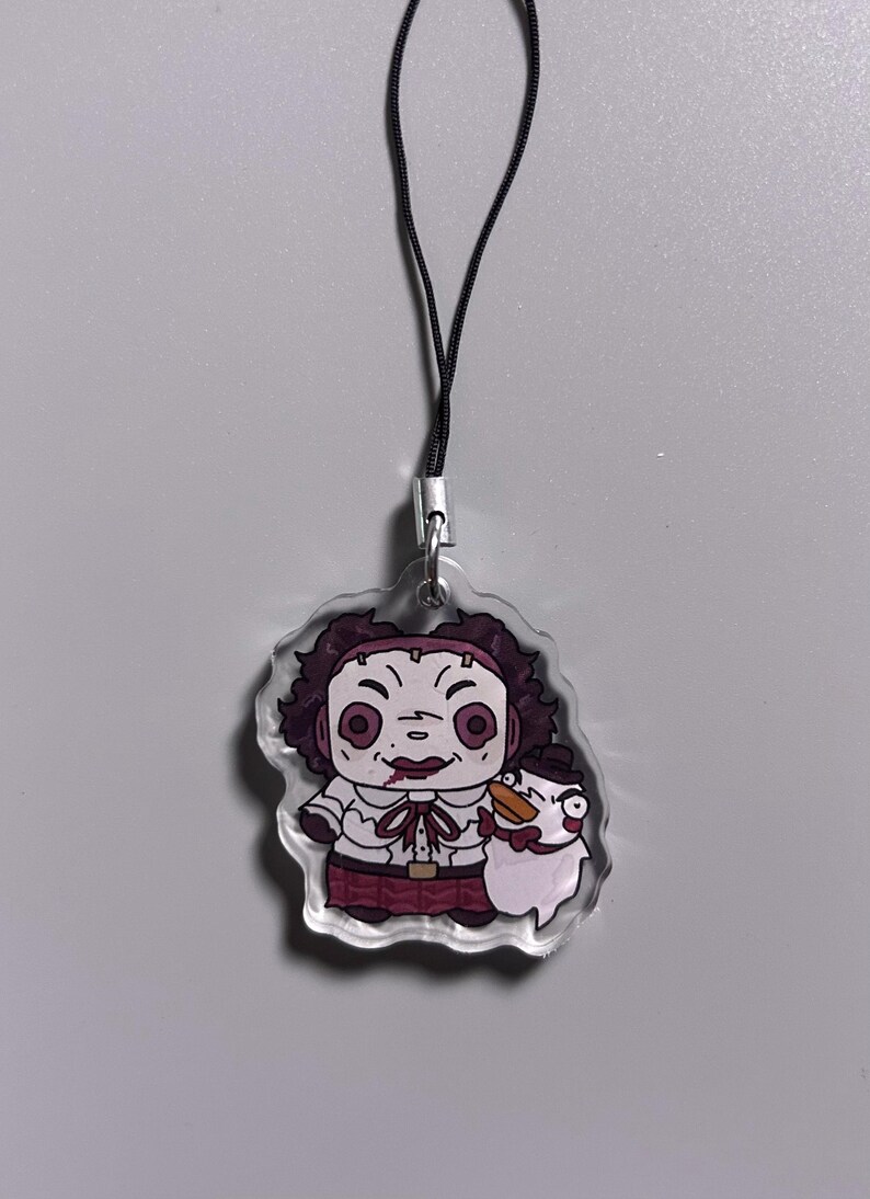May include: A clear acrylic charm featuring a cartoon character with a chainsaw and a snowman. The character has dark curly hair, a white shirt, and a red skirt. The charm is attached to a black cord.