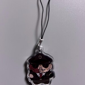 May include: A clear acrylic charm featuring a cartoon illustration of a man in a police uniform, wearing a hat and sunglasses, with a cigarette in his mouth. The charm has a black cord attached for hanging.