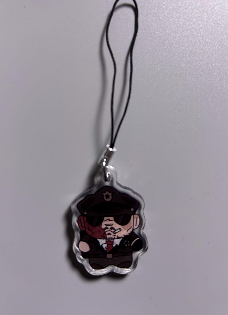May include: A clear acrylic charm featuring a cartoon illustration of a man in a police uniform, wearing a hat and sunglasses, with a cigarette in his mouth. The charm has a black cord attached for hanging.