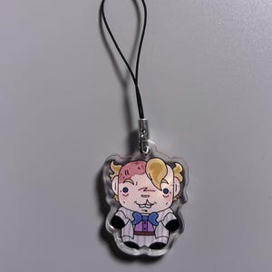 May include: A clear acrylic charm featuring a cartoon character with blonde hair, a purple bow tie, and a striped suit. The charm is attached to a black lanyard with a silver clasp. The character has a unique facial expression.