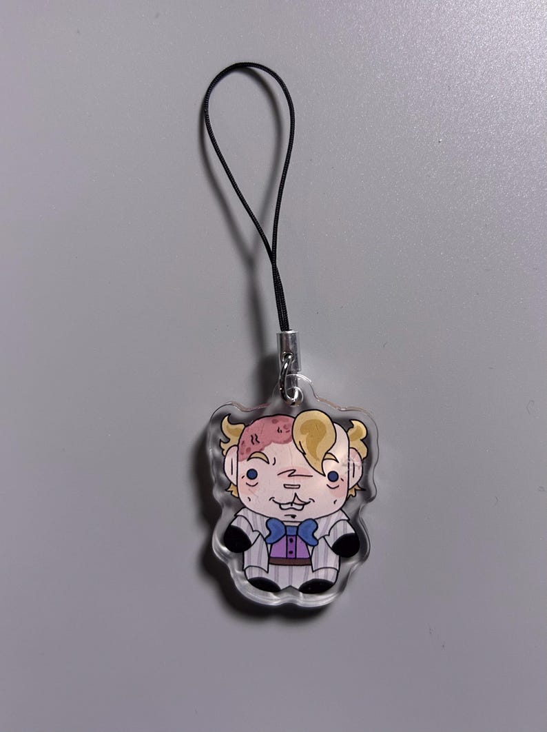 May include: A clear acrylic charm featuring a cartoon character with blonde hair, a purple bow tie, and a striped suit. The charm is attached to a black lanyard with a silver clasp. The character has a unique facial expression.