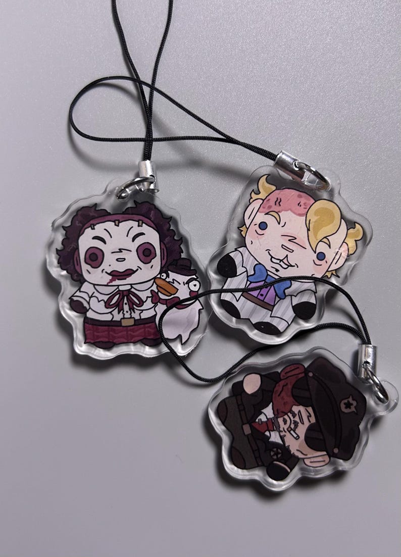 May include: Three clear acrylic charms with cartoon illustrations of characters. One charm features a character in a white shirt and maroon skirt, holding a duck. Another charm depicts a character in a suit. The third charm shows a character in a dark coat.