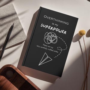 May include: A black hardcover notebook featuring white text that reads OVERTHINKING is my SUPERPOWER and These are my very carefully crafted notes. The cover design includes a white line drawing of a tangled scribble and a paper airplane.