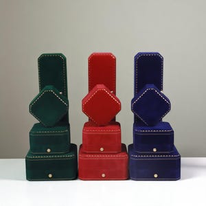 May include: A collection of velvet jewelry boxes in green, red, and blue. Each set includes boxes of varying sizes, with gold stitching and a small button closure. The boxes are arranged in a stacked display, showcasing their different shapes and sizes.