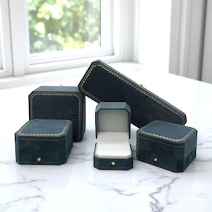 Puede incluir: Una colección de joyeros de terciopelo verde azulado en varias formas y tamaños. Las cajas tienen ribetes dorados y pequeños pomos dorados. Una caja está abierta, revelando un interior blanco.