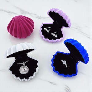 Op de afbeelding: Een verzameling schelpvormige sieradendoosjes in verschillende kleuren: fuchsia, lavendel, wit en blauw. Elke doos is open en toont een sieraad: oorbellen, een ketting en een ring. De doosjes staan op een wit marmeren oppervlak.