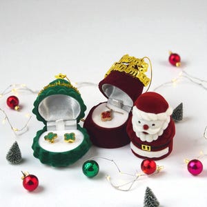 以下が含まれることがあります： クリスマスをテーマにした画像。ジュエリーボックスと装飾品が写っています。緑色のベルベットの箱には金のイヤリング、赤い箱にはネックレスが入っています。サンタクロースの置物と赤と緑のオーナメントが、お祝いのシーンを完成させています。