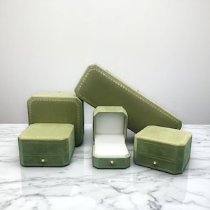 Puede incluir: Una colección de joyeros de terciopelo verde oliva de varias formas y tamaños. Los joyeros tienen detalles dorados y un interior blanco. Están colocados sobre una superficie de mármol.