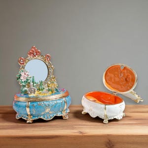 Könnte beinhalten: Zwei dekorative Schmuckkästchen. Eines ist blau mit goldenen Akzenten, einem Spiegel und Miniatur-Parfümflaschen. Das andere ist weiß mit goldenen Verzierungen und einem orangefarbenen Innenraum. Beide sind verziert und stehen auf einer Holzoberfläche.