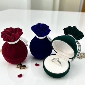 以下が含まれることがあります： 赤、青、緑のベルベットリングボックス3つ。緑色のボックスは開いており、透明な石が付いたシルバーリングが見えます。各ボックスには、白いコードが付いた巾着型の留め具が付いています。小さなハート型の装飾が散らばっています。