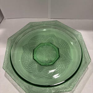 May include: A light green, octagonal glass plate featuring a central octagonal medallion and an intricate, textured geometric pattern radiating outward toward the rim. The glass has a translucent quality with a distinct pressed design.