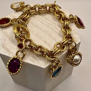 Puede incluir: Una pulsera de dijes dorada con un diseño de cadena texturizada. La pulsera presenta varios dijes con piedras preciosas de colores en rojo, morado y azul, y un dije de perla. La pulsera se exhibe sobre una superficie blanca.