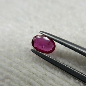 Può includere: Una gemma di rubino ovale e sfaccettata tenuta da una pinzetta d'argento. Il rubino è di un colore rosso intenso e ricco con una leggera lucentezza. Lo sfondo è un tessuto bianco morbido e strutturato.
