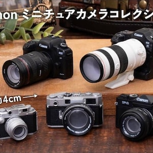Op de afbeelding: Een verzameling van vijf miniatuur Canon cameramodellen, waaronder twee spiegelreflexcamera's met zwarte en witte lenzen en drie compactcamera's in zwarte en zilveren afwerkingen. Een label geeft een schaal van ongeveer 4 cm aan.