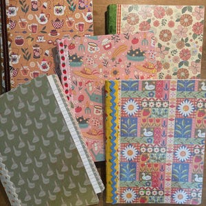 May include: Four rectangular notebooks with decorative covers. One features tea-themed illustrations, one has a floral pattern, one displays a garden motif with strawberries, and one shows a grid of flowers, leaves, and white ducks with a blue trim.