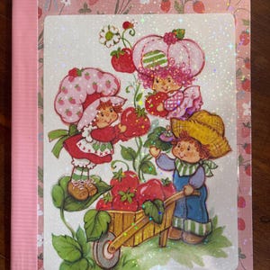May include: A rectangular notebook with a pink strawberry-patterned cover featuring a central holographic illustration of three characters harvesting strawberries into a wooden wheelbarrow, surrounded by green leaves and small white flowers.