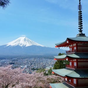 Può includere: Una vista panoramica del Monte Fuji in Giappone, con una tradizionale pagoda rossa e bianca in primo piano. I fiori di ciliegio incorniciano la scena e un cielo azzurro fa da sfondo alla montagna innevata e alla città sottostante.