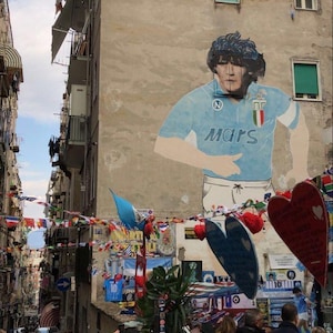 Puede incluir: Escena callejera en Nápoles, Italia, con un mural de Diego Maradona con una camiseta de fútbol azul claro. Banderas de varias naciones cuelgan sobre la calle. La gente camina por la estrecha calle.