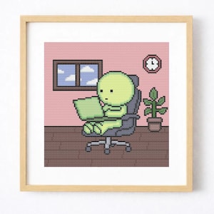 May include: A framed cross-stitch artwork depicts a green figure seated at a desk, working on a laptop. The figure is in a gray office chair. A window, clock, and potted plant are also in the room. The background is pink.
