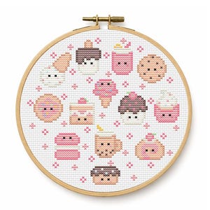 May include: A circular wooden embroidery hoop containing white fabric with a cross-stitch pattern of various colorful desserts including ice cream cones, cookies, cupcakes, and macarons, all featuring small black cross-stitched eyes.