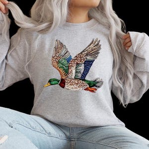 May include: A gray sweatshirt featuring a detailed, colorful embroidered duck in flight. The duck has green, blue, brown, and red accents. The sweatshirt is a casual style, suitable for everyday wear.