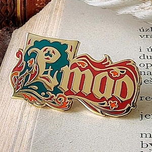 Può includere: Una spilla smaltata color oro con la parola "mao" in rosso, bordata d'oro, e un design turchese e oro. La spilla è su una pagina di libro con una piuma e una pila di libri sullo sfondo.