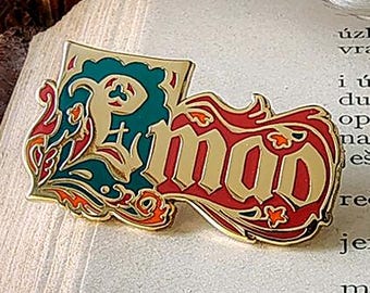 Puede incluir: Un pin de esmalte dorado con la palabra "mao" en rojo, contorneada en dorado, y un diseño turquesa y dorado. El pin está sobre una página de libro con una pluma y una pila de libros en el fondo.