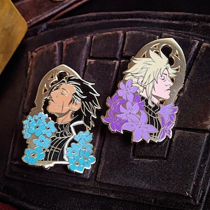 May include: Two gold-colored enamel pins featuring anime-style character profiles. One pin has a character with black hair and blue flowers, the other has a character with blonde hair and purple flowers. Both pins have a decorative gold border.