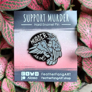 Puede incluir: Un pin de esmalte duro con el texto "SUPPORT MURDER" y "MURDER is ok" en un diseño de cuervo negro y gris. El pin está sobre una tarjeta con un fondo degradado rosa y gris, rodeado de hojas rosas y verdes.