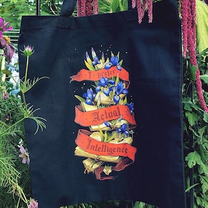 Op de afbeelding: Een zwarte draagtas met een grafische print van open boeken verweven met blauwe irisbloemen en een rood lintbanner. De tekst op de banner luidt I prefer Actual Intelligence in een gotische letterstijl, gecentreerd op de voorzijde van de tas.