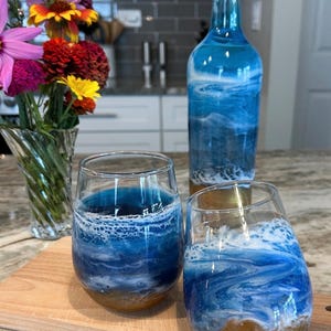 K&ouml;nnte beinhalten: Eine Glasflasche und zwei stiellose Weingl&auml;ser mit einem blau-wei&szlig;en Meereswellen-Design und einem sandbraunen Boden. Die Flasche tr&auml;gt den Text Beth Scotts Studio. Eine klare Glasvase mit bunten Blumen ist ebenfalls zu sehen.