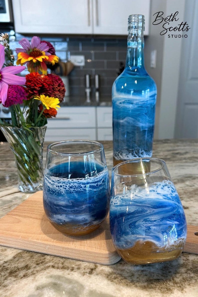 K&ouml;nnte beinhalten: Eine Glasflasche und zwei stiellose Weingl&auml;ser mit einem blau-wei&szlig;en Meereswellen-Design und einem sandbraunen Boden. Die Flasche tr&auml;gt den Text Beth Scotts Studio. Eine klare Glasvase mit bunten Blumen ist ebenfalls zu sehen.