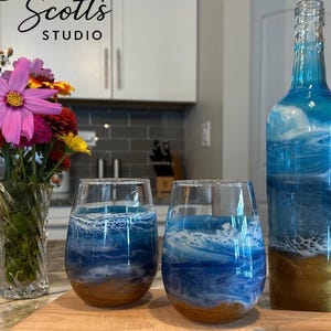 K&ouml;nnte beinhalten: Eine Glasflasche und zwei stiellose Weingl&auml;ser mit einem blau-wei&szlig;en Ozeanwellen-Harzdesign auf goldfarbenem Grund. Eine durchsichtige Glasvase mit einem Strau&szlig; aus rosa, orangefarbenen und gelben Blumen steht links neben den Glaswaren.