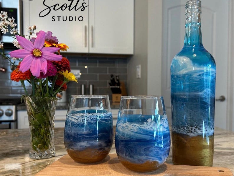 K&ouml;nnte beinhalten: Eine Glasflasche und zwei stiellose Weingl&auml;ser mit einem blau-wei&szlig;en Ozeanwellen-Harzdesign auf goldfarbenem Grund. Eine durchsichtige Glasvase mit einem Strau&szlig; aus rosa, orangefarbenen und gelben Blumen steht links neben den Glaswaren.