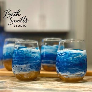 K&ouml;nnte beinhalten: Vier stiellose Weingl&auml;ser mit einem dekorativen Harzdesign, das eine Meereslandschaft mit sandgoldener Basis, wirbelndem blauem Wasser und wei&szlig;em Meeresschaum nachahmt. Der Text Beth Scotts Studio ist oben links in Schwarz aufgedruckt.