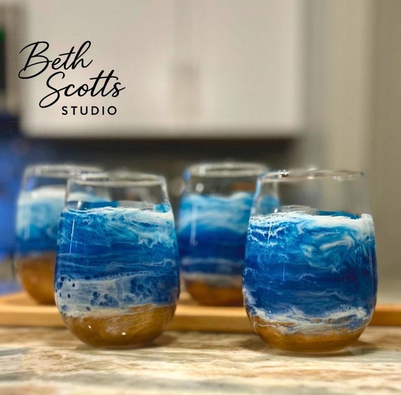K&ouml;nnte beinhalten: Vier stiellose Weingl&auml;ser mit einem dekorativen Harzdesign, das eine Meereslandschaft mit sandgoldener Basis, wirbelndem blauem Wasser und wei&szlig;em Meeresschaum nachahmt. Der Text Beth Scotts Studio ist oben links in Schwarz aufgedruckt.