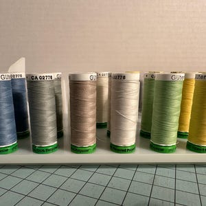 May include: A collection of spools of Gütermann thread in various colors, including blue, gray, beige, white, green, and yellow. The spools are arranged on a white tray, with the thread visible. Each spool has a green base and a label.