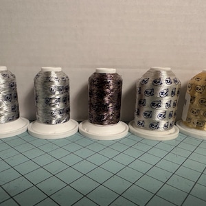 May include: Five spools of metallic thread in silver, dark brown, and gold, each on a white base. The silver spools have a repeating blue logo pattern. The gold spool has a gold logo pattern.