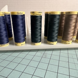 May include: A collection of spools of thread in various colors, including navy blue, dark green, and tan. The spools are arranged on a white plastic organizer. Each spool has a yellow label with text.