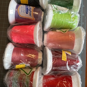 May include: A collection of spools of nylon thread in various colors, including green, purple, red, and pink. The spools are wrapped in clear plastic and have white bases. Some spools have price tags attached. The text "Nylon Thread" is visible.