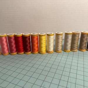 May include: A selection of sewing thread spools in a range of colours, from pinks and reds to yellows and browns. The spools are lined up, showing their labels. The background is a plain, light colour.