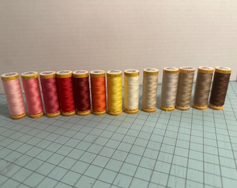 May include: A collection of spools of thread in various colors, including shades of pink, red, orange, yellow, beige, and brown. The thread spools are arranged in a row, with the labels facing the viewer. The background is a light color.