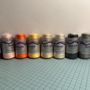 May include: A row of eight spools of SureLock thread in various colors, including burgundy, pink, orange, yellow, cream, beige, dark gray, light gray, and white. Each spool is labeled with the SureLock brand name.