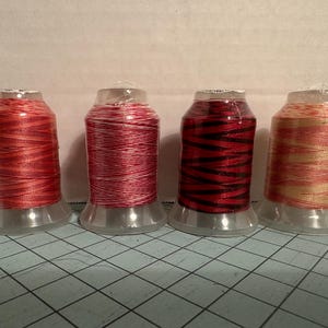 May include: Four spools of thread in shades of red, pink, and gold. The thread is wound around clear plastic spools. One spool has black stripes. The spools are on a blue grid surface.