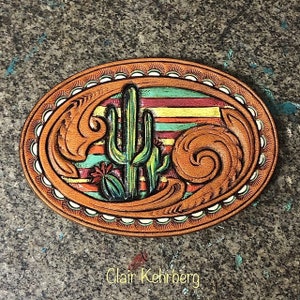 May include: A leather belt buckle with a colorful design featuring a cactus and a striped background. The buckle is oval shaped and has a decorative border.