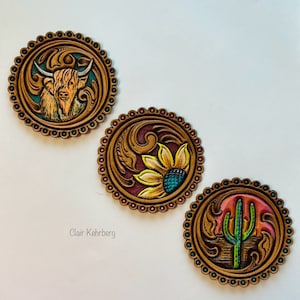 May include: Three round leather coasters with intricate designs. The first coaster features a brown and white Highland cow, the second features a yellow sunflower with turquoise accents, and the third features a green cactus with a pink sunset.